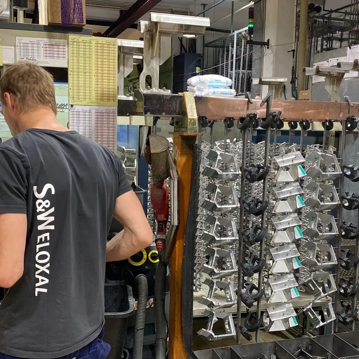 Mitarbeiter im grauen T-Shirt mit S&W-Eloxal-Schriftzug am Rücken arbeitet an einem Gestell voller Aluminiumteile