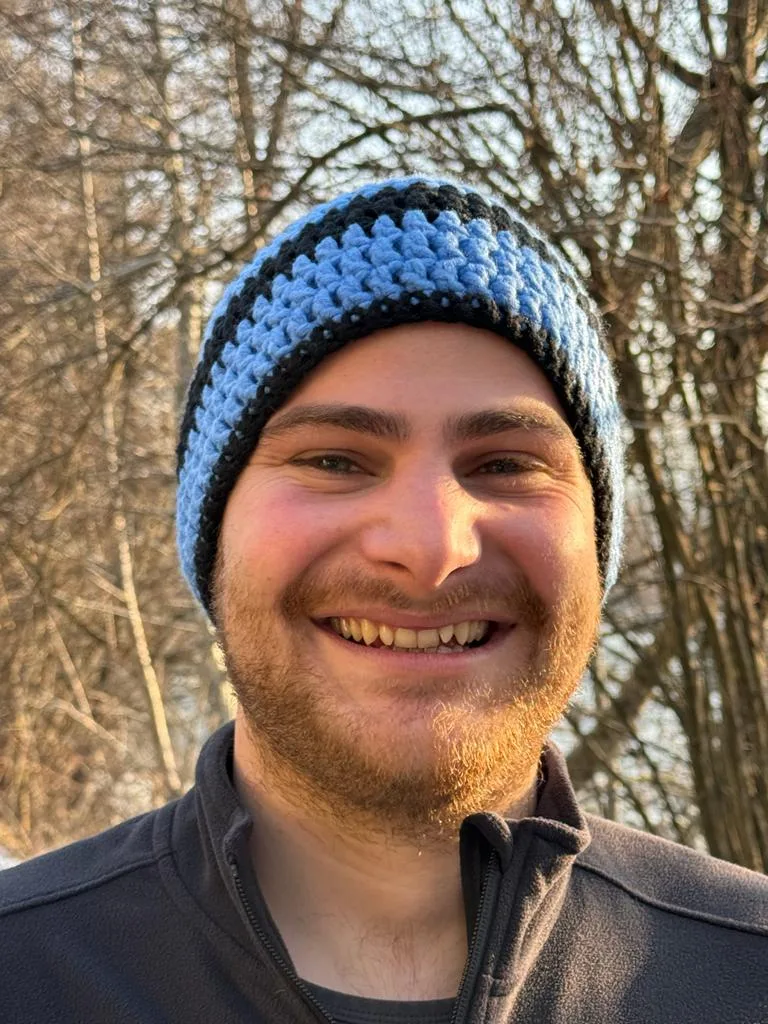 Daniel Wallenböck mit blau-schwarzer Strickmütze lächelt im Freien vor kahlen Bäumen